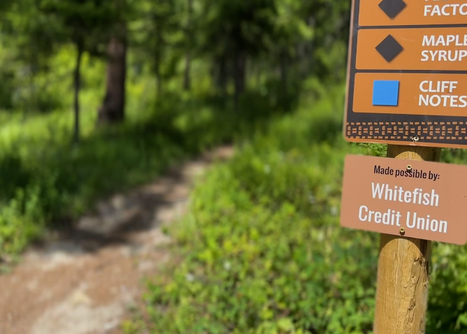 A wooden trail sign along a forest path credits Whitefish Credit Union for supporting the site.