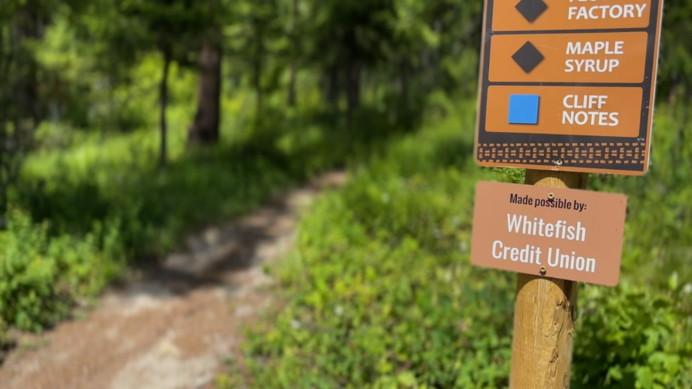 A wooden trail sign along a forest path credits Whitefish Credit Union for supporting the site.